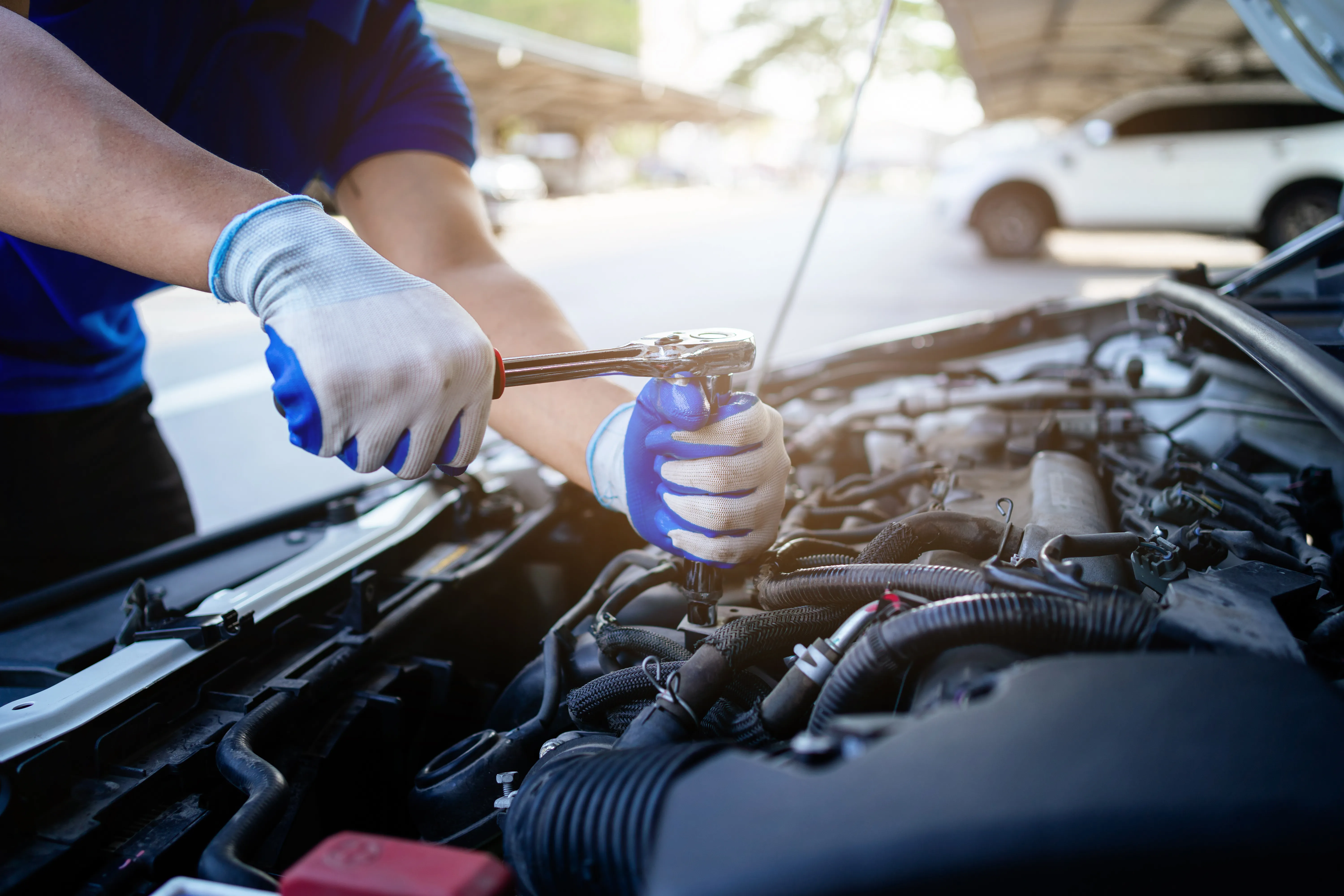 A man is working on a car engine. He is wearing blue gloves and a blue shirt. The car is parked on the side of the road