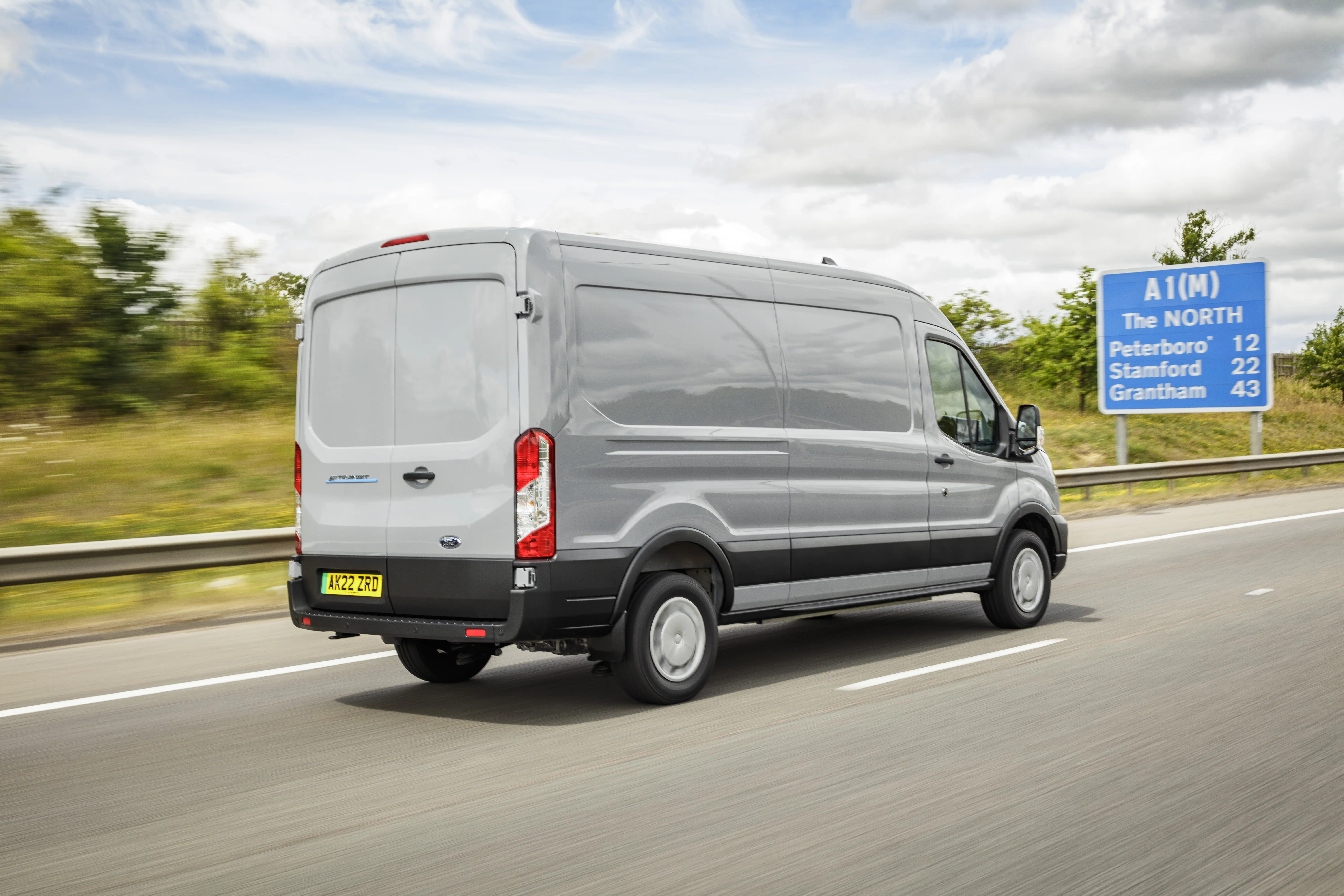 A silver Ford e-van drives along a road with a blue road sign in background
