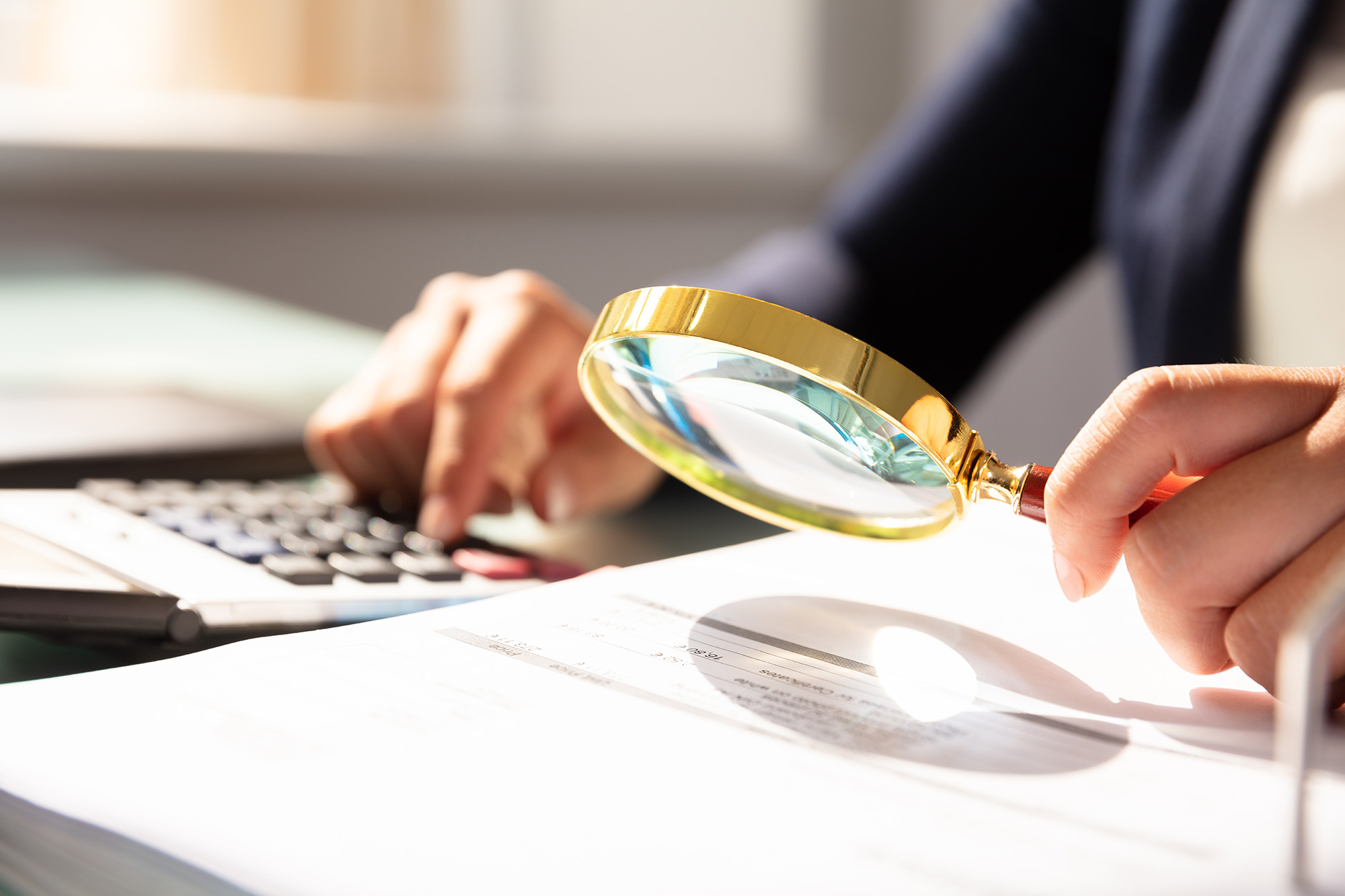 Businessman sitting at a desk with papers and keyboard holding a magnifying glass